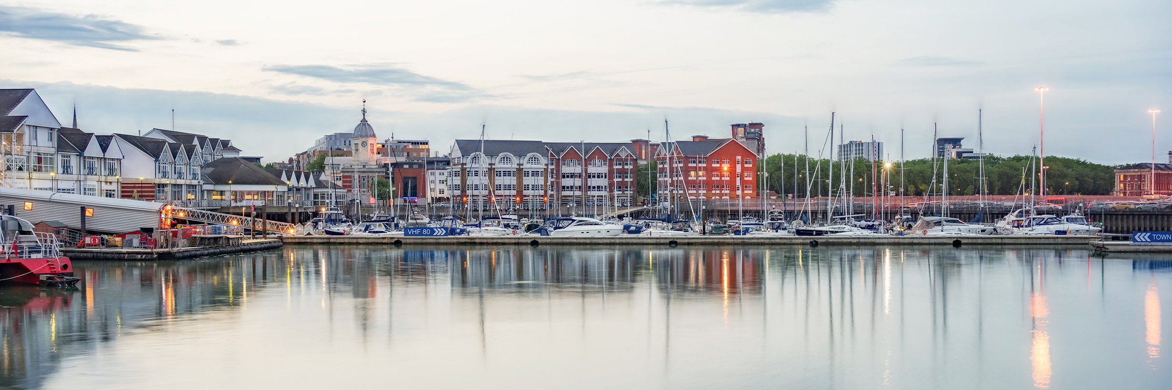 Moored yachts on still water at dusk, with the city waterfront behind them